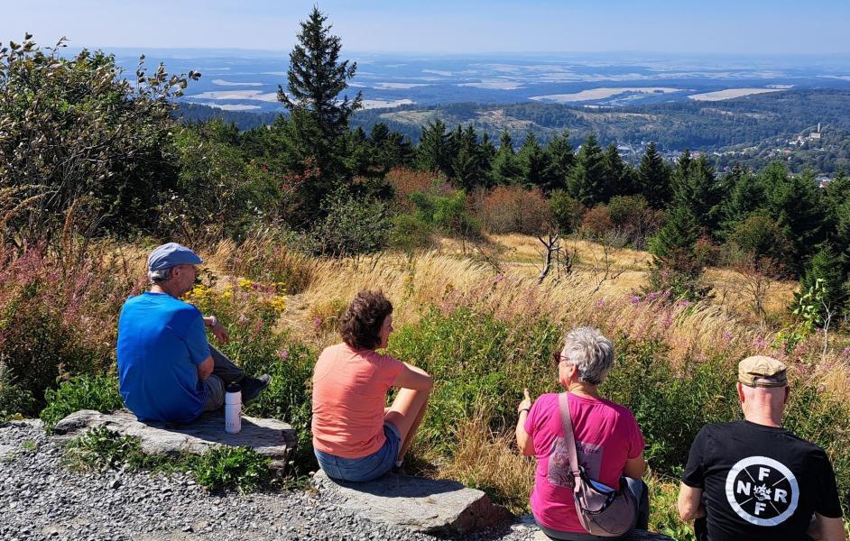 Vier Personen sitzen mit dem Rück zur Kamera auf dem Boden. Blick ins Tal bei blauem Himmel. 