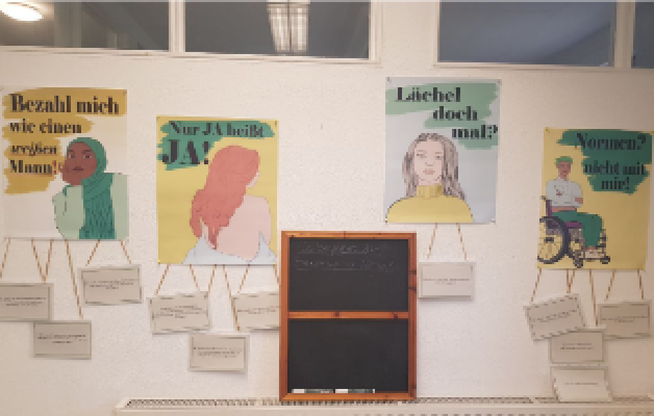 Vier Plakate hängen an einer weißen Wand. Auf den Plakaten steht: (1) Bezahl mich wie einen weißen Mann!; (2) Nur Ja heißt Ja!; (3) Lächel doch Mal?; (4) Normen? Nicht mit mir!; In der Mitte steht eine Tafel, darauf steht: Wo begegnet dir Sexismus im Alltag?