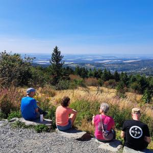 Vier Personen sitzen mit dem Rück zur Kamera auf dem Boden. Blick ins Tal bei blauem Himmel. 